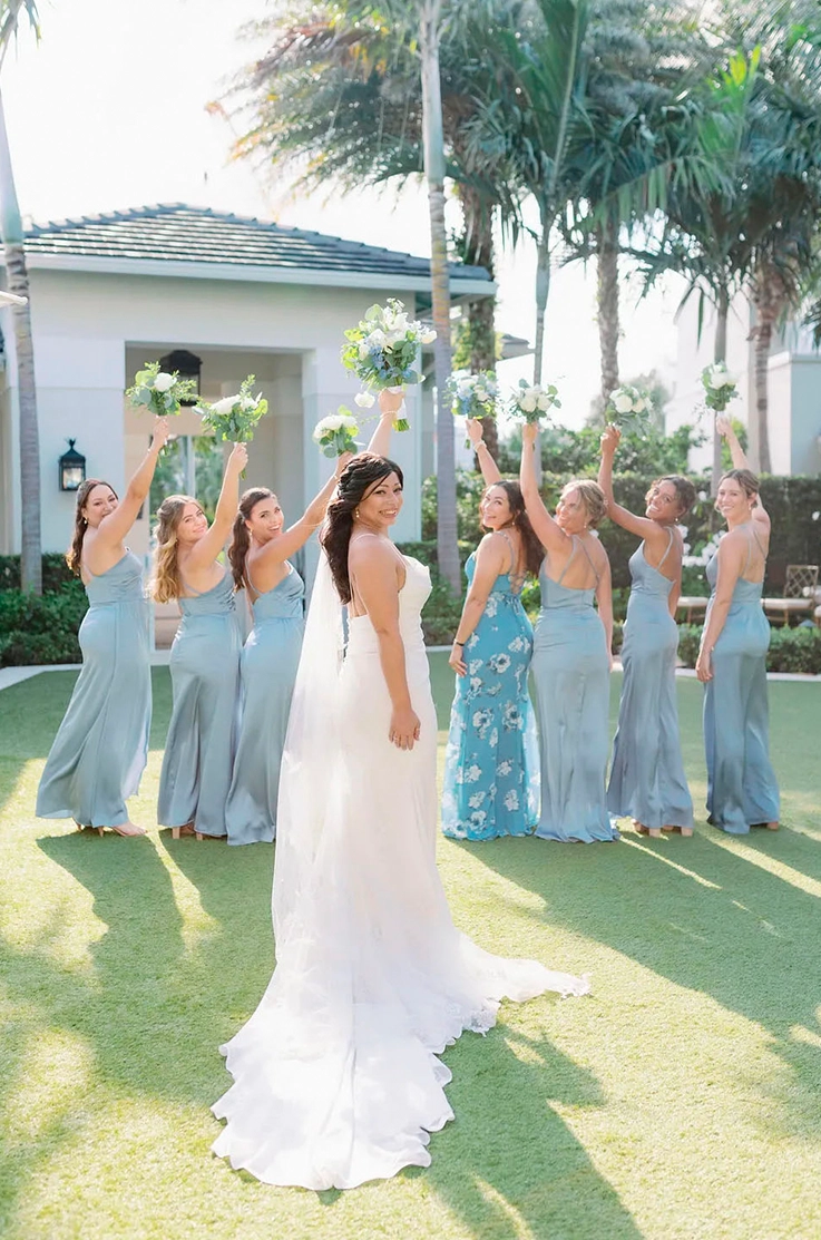 Bride in a crepe pleated gown with fit and flare skirt and a veil, raises a bouquet in the middle of her bridesmaids.