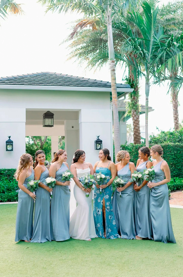 Bride in a crepe pleated bodice gown stands in the middle of bridesmaids in blue dresses, in front of palm trees.