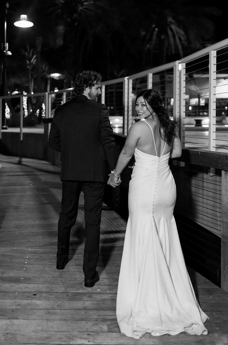 Bride looks over her shoulder in a low back fit and flare gown with buttons, is walking beside her groom in black and white.