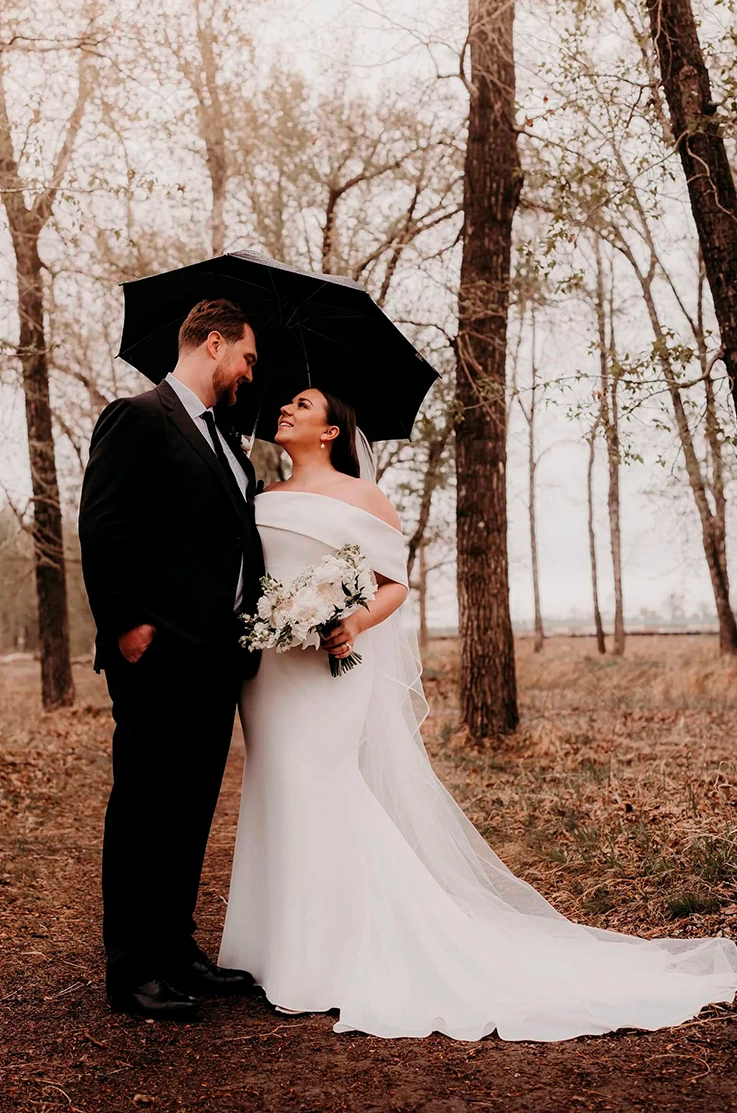 Bride in a crepe fit and flare gown with a portrait collar and veil, poses with groom under an umbrella in a forest.