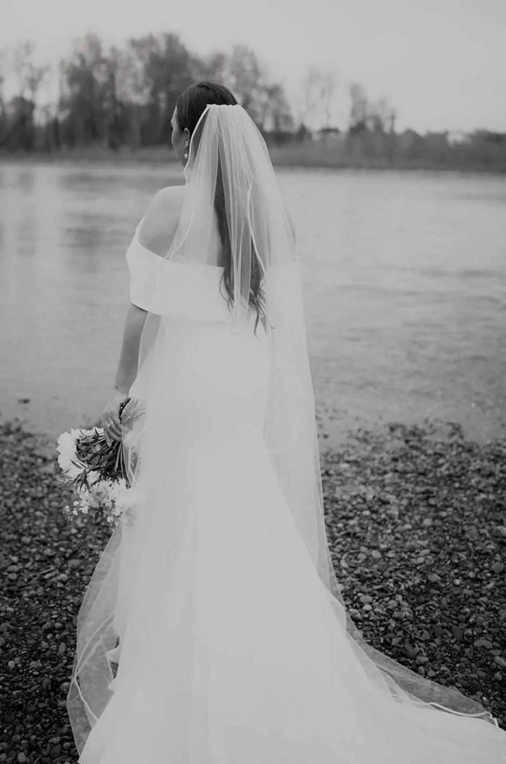 Bride in an off-the-shoulder crepe wedding dress with a veil, is standing at a lakeside in black and white.