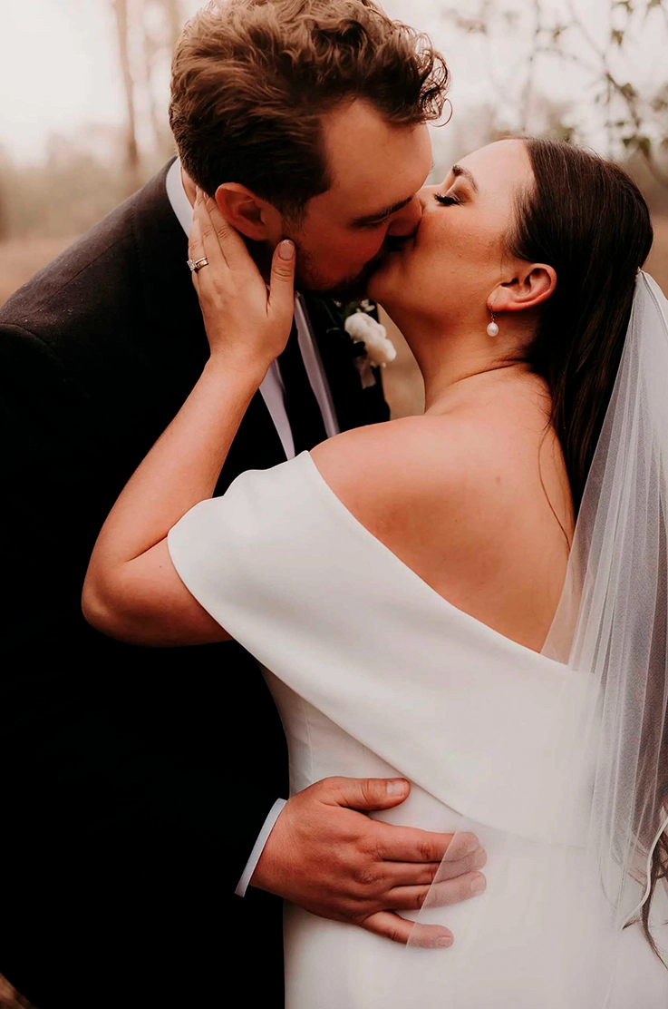 Close-up of groom kissing bride in an off-the-shoulder crepe gown with a veil in a rainy forest setting.