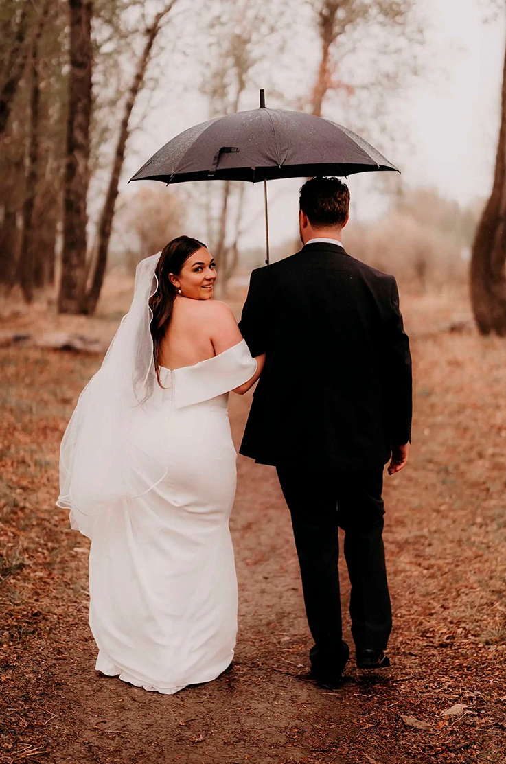 Bride in a fit and flare crepe gown with a portrait collar and veil, poses and walks with groom under an umbrella in fall leaves.