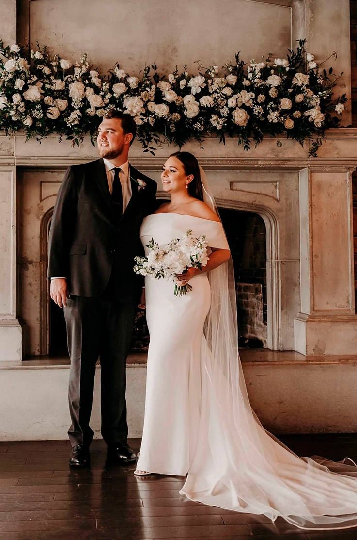 Bride in a portrait collar fitted crepe gown with a long veil, is holding a bouquet next to groom by a white stone fireplace. 