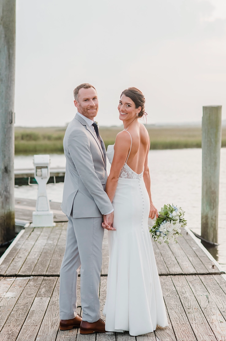 Bride in a backless lace and crepe gown with a buttoned fit and flare skirt, looks back with groom on a waterfront dock.