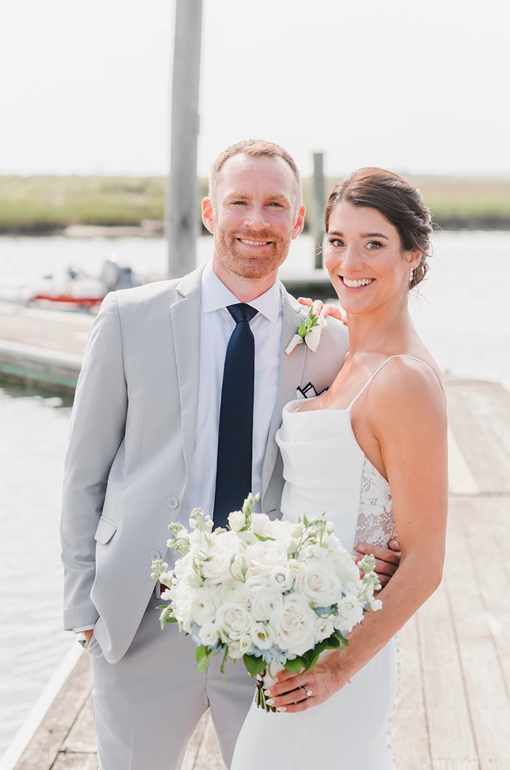 Bride in a scoop neck crepe wedding dress with floral lace detail, is holding a white bouquet on a dock and poses with groom.