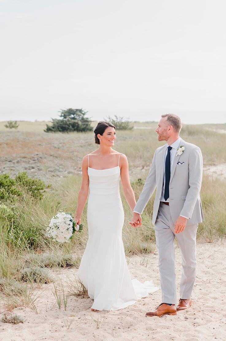 Bride in a pleated bodice gown with a crepe fit and flare skirt and a scoop neck, walks alongside her groom on a beach. 