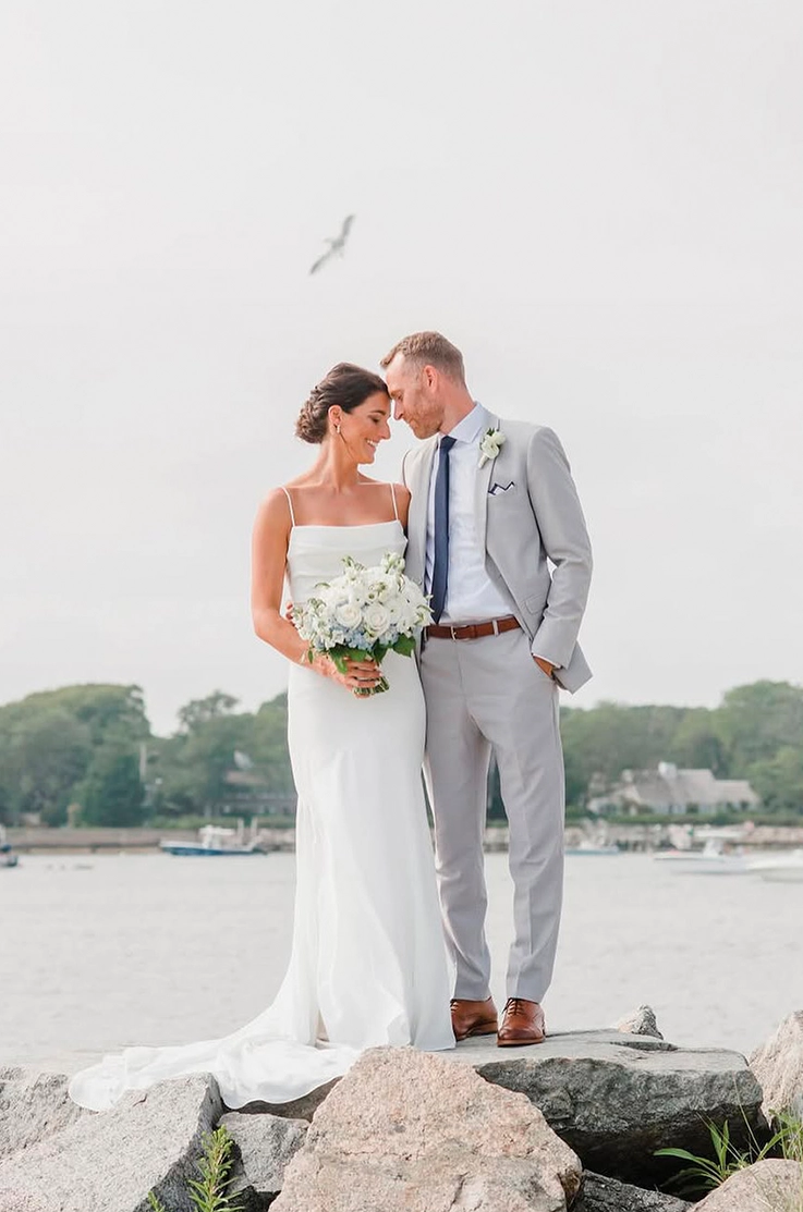Bride in a crepe fit and flare gown with a scoop neckline and straps, is holding a bouquet next to her groom on a rocky shore.