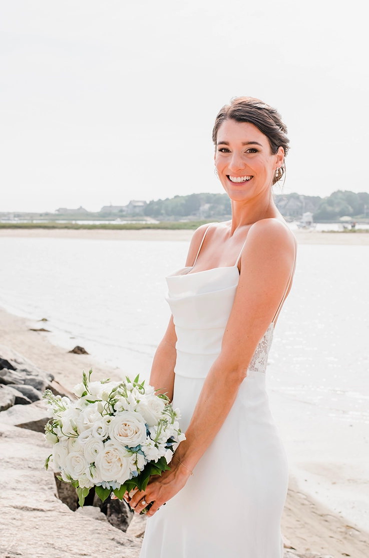 Close-up of bride in a pleated crepe bodice gown with a floral lace back and a scoop neck, holding a bouquet on a shoreline.Close-up of bride in a pleated crepe bodice gown with a floral lace back and a scoop neck, holding a bouquet on a shoreline.