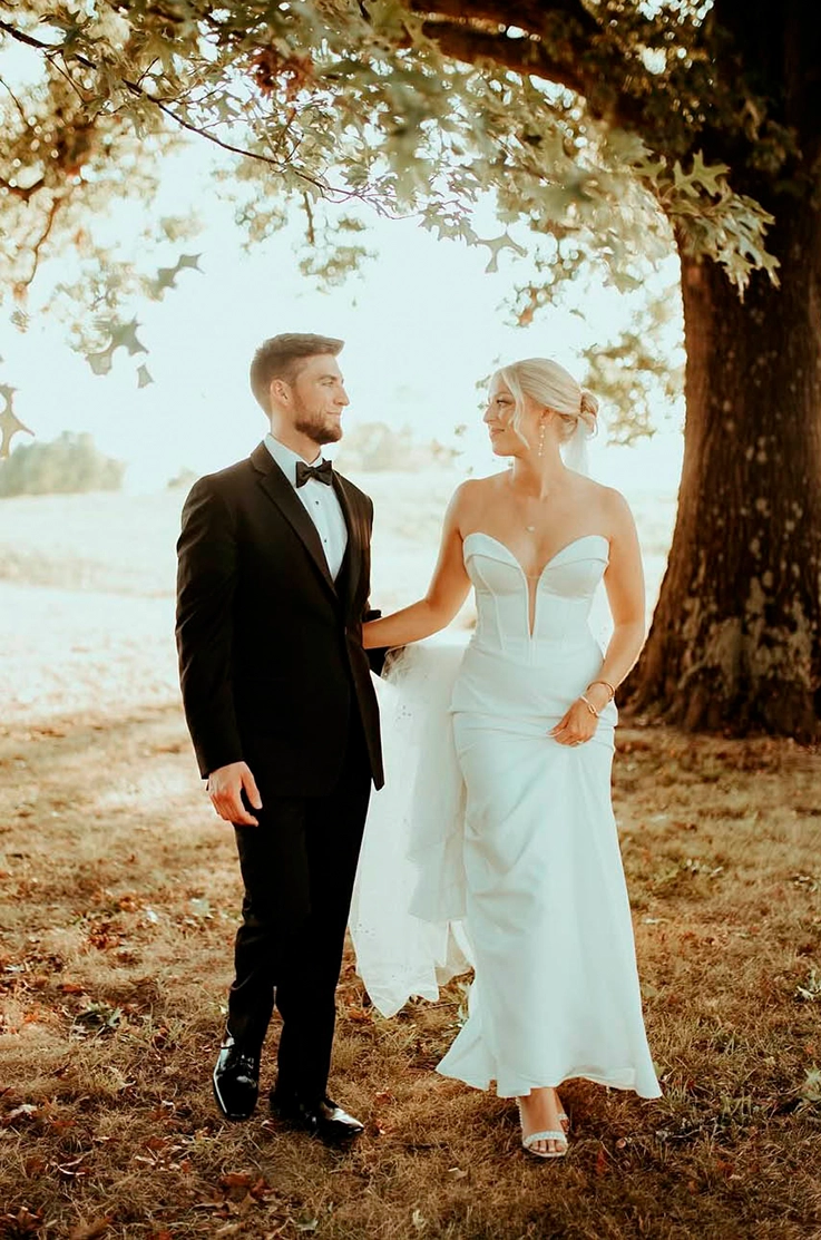 Bride in a strapless crepe wedding dress with a sweetheart neckline, walks alongside her groom under a large sunlit tree.