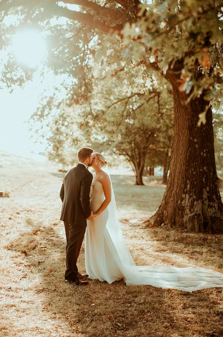 Groom kisses his bride in a crepe fit and flare gown with a long veil, beneath golden oak trees in the sunlight. 