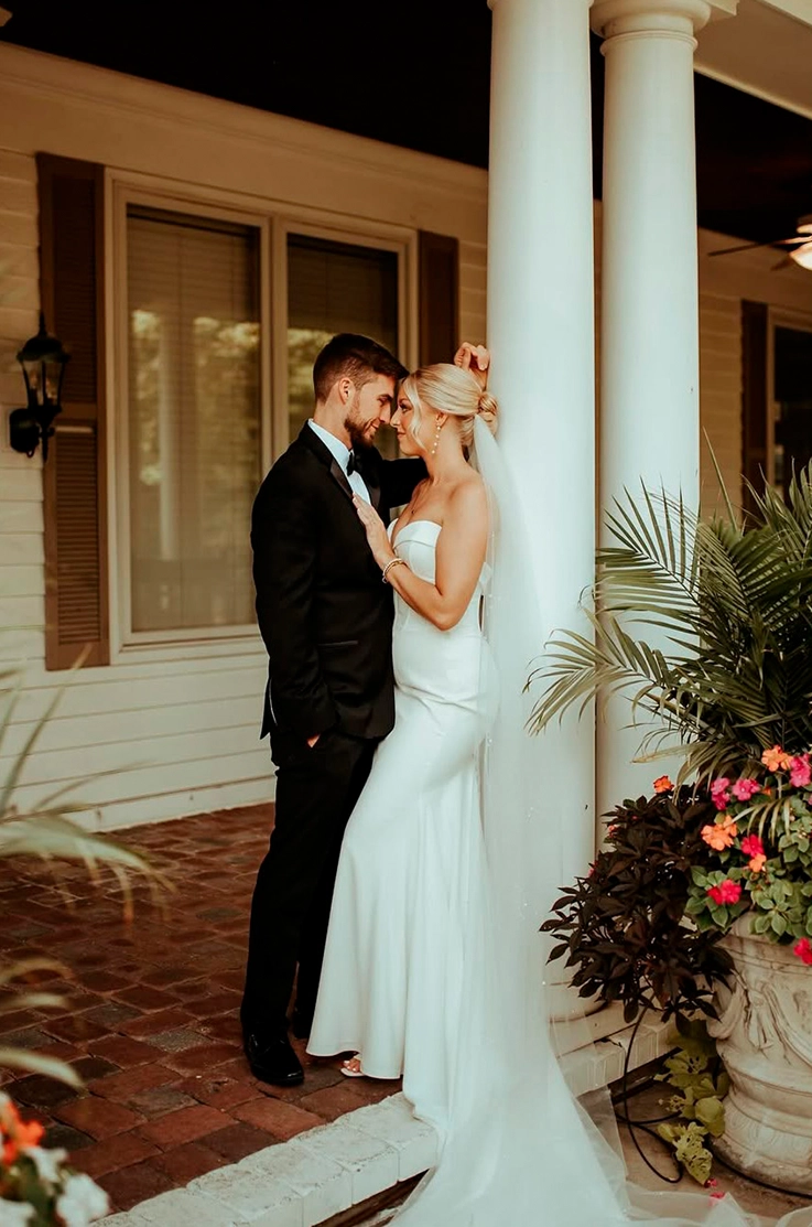 Bride in a fit and flare crepe gown with a sweetheart neck and a veil, faces her groom on a veranda with pillars and flowers. 