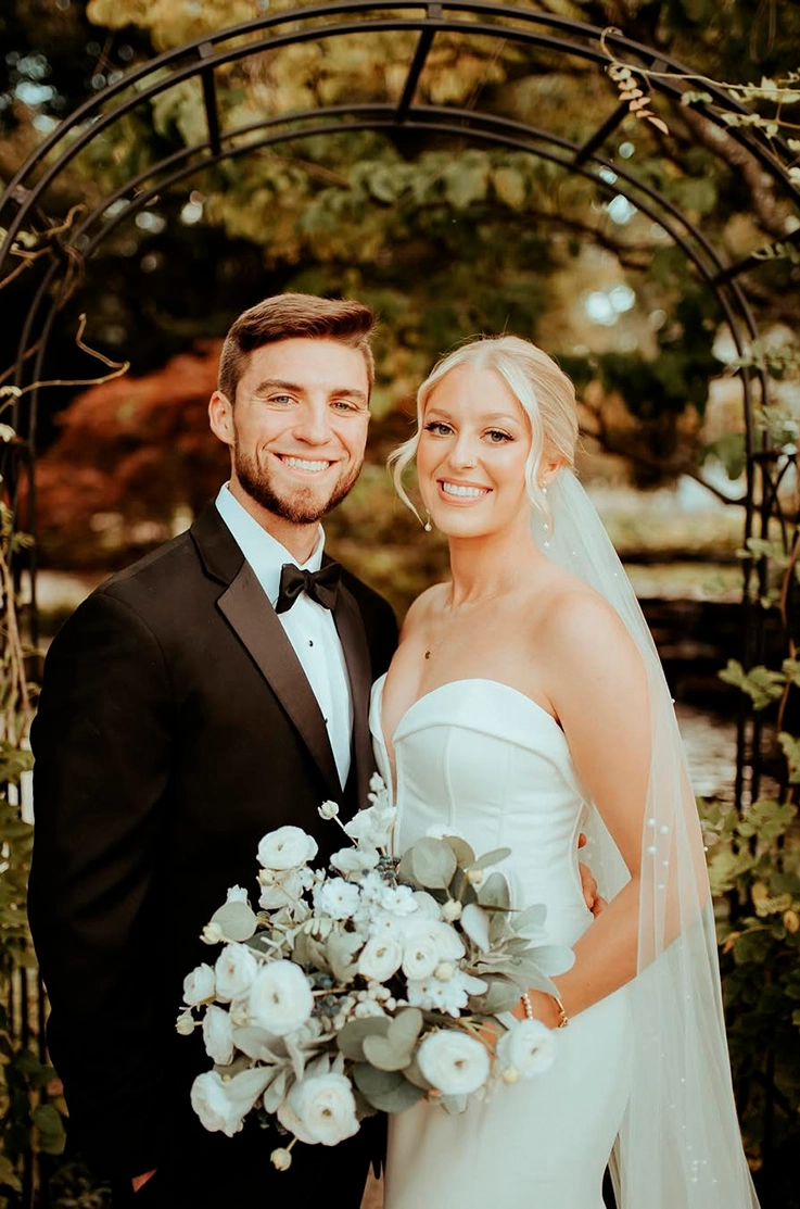 Bride in a fitted crepe gown with a plunging sweetheart neckline and a veil, poses next to groom under an iron garden arch.