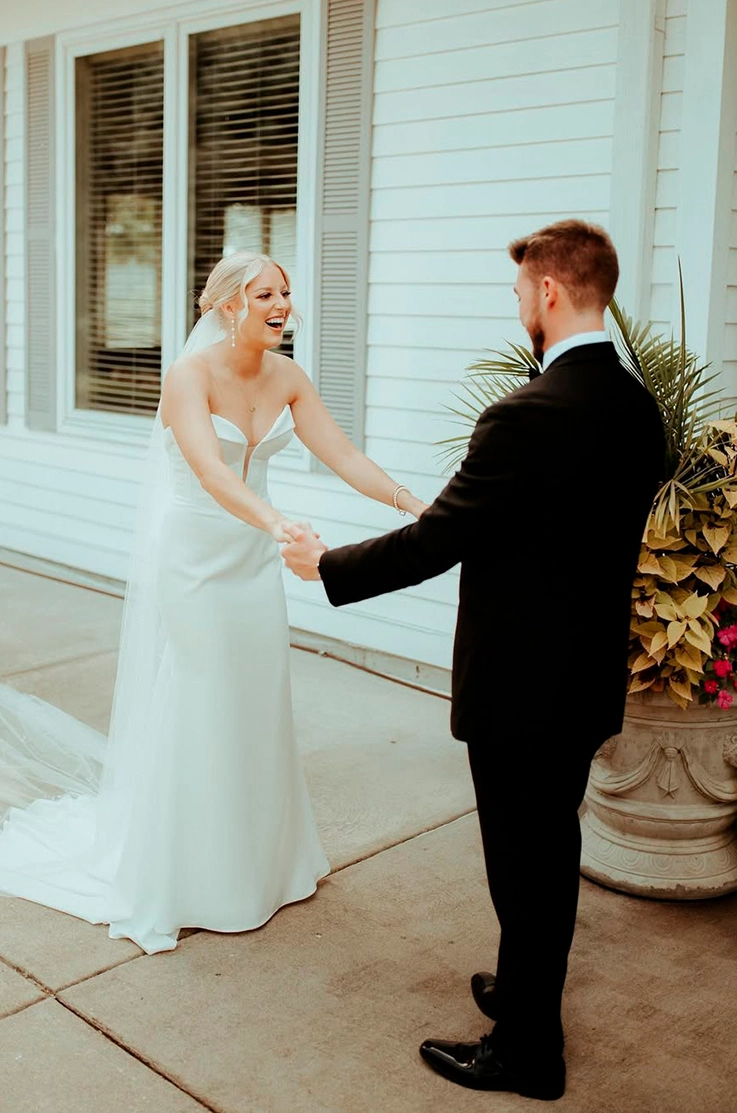 Groom faces bride in a strapless sweetheart neck gown with corset seam detail on the bodice and a veil, outside the venue. 