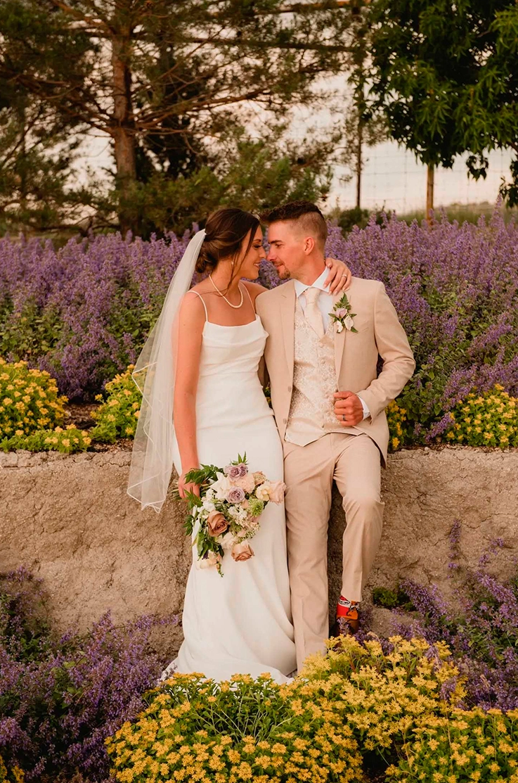 Bride in a fit and flare crepe wedding dress, poses beside groom and holds a bouquet, sitting on rocks in a serene garden.