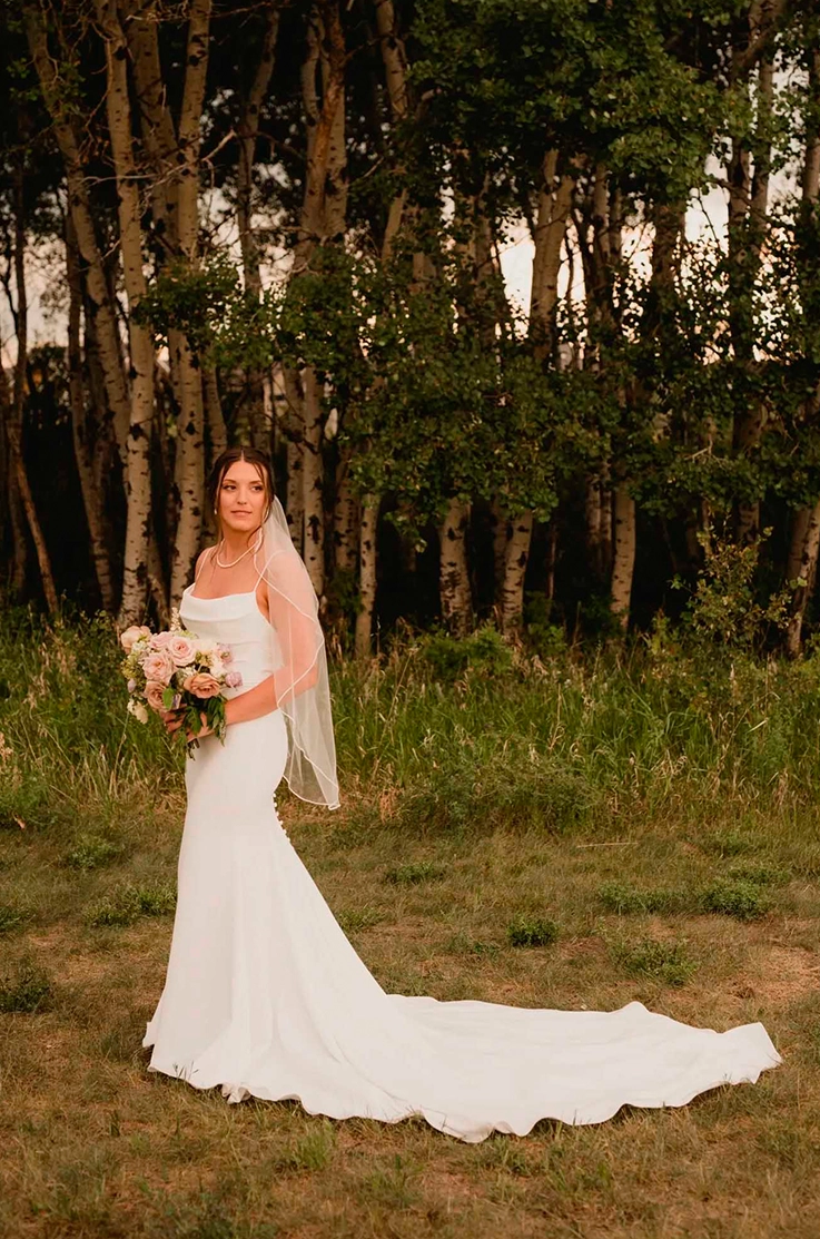 Bride poses in a scoop neck crepe gown with fit and flare skirt and a veil, in a forest setting, holding a blush bouquet.