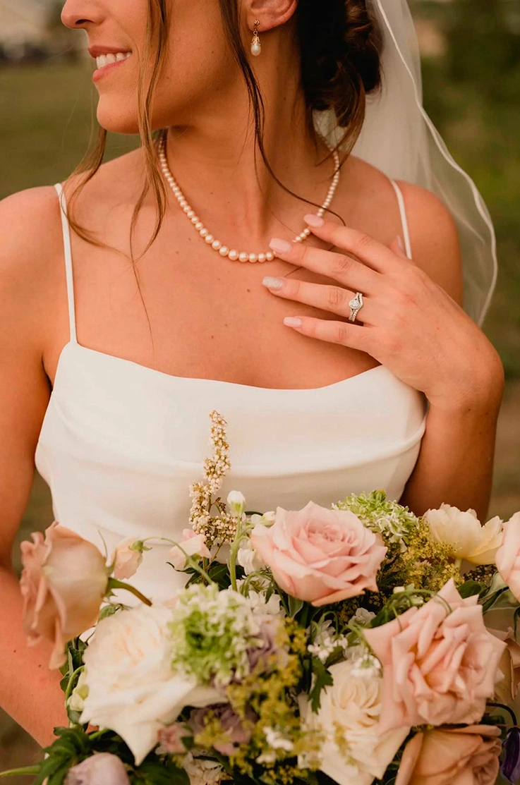 Close-up of bride in a pleated crepe bodice gown with thin straps and a veil, is holding a pink and white bouquet outdoors. 