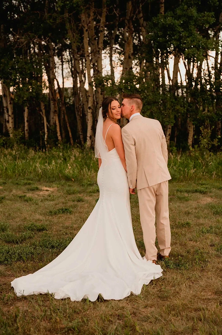 Bride in a fit and flare sleeveless gown with buttons and a veil, poses next to groom in front of trees in an open field.