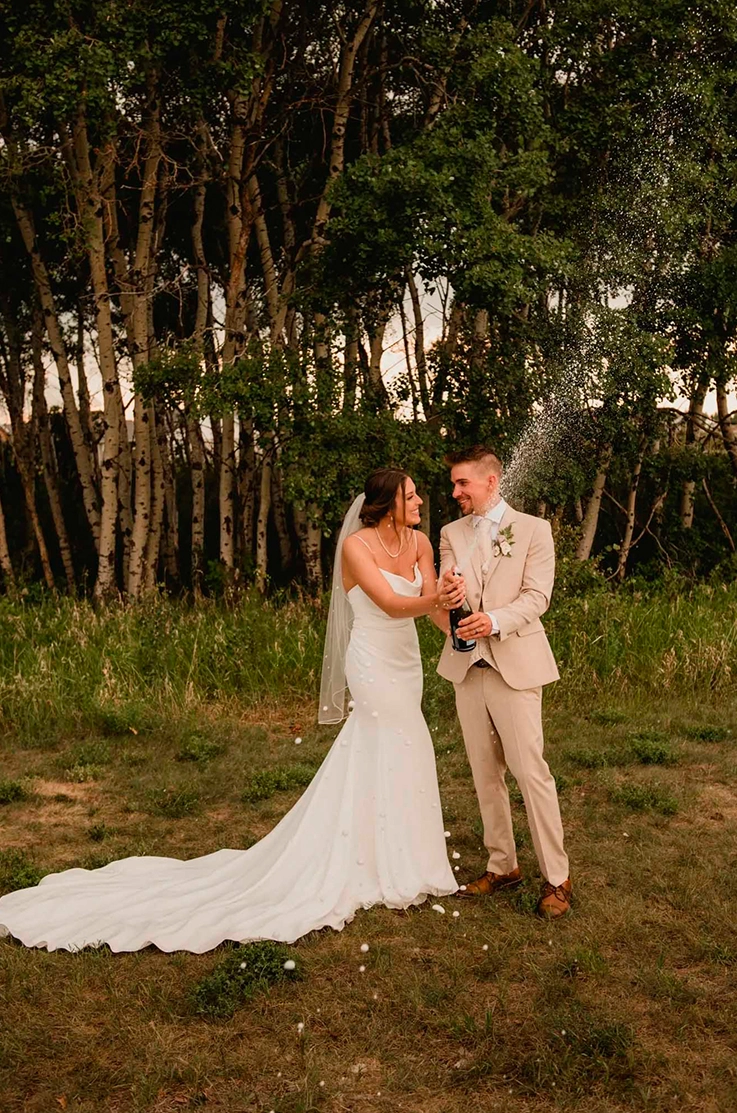 Bride in a draped crepe fitted gown with a scoop neckline and veil, stands next to groom and they pop open champagne outdoors. 