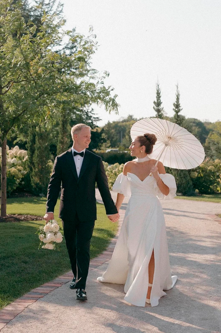 Bride in an off-shoulder puff sleeve gown in satin faille, is holding an umbrella and walks beside her groom on a garden path.
