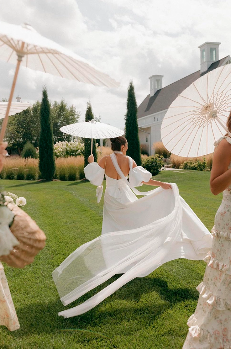 Bride in a flowing satin faille wedding dress with puff sleeves and open back, is walking through manicured estate gardens.