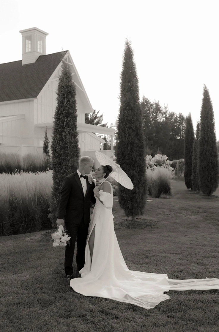 Bride in a satin faille gown with a skirt slit, scarf and separate sleeves, poses with groom outside venue in black and white.