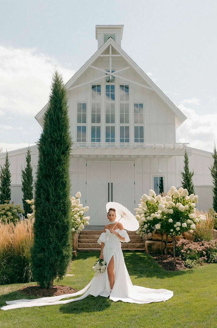 Bride in an off-shoulder puff sleeve gown with a skirt slit and scarf, is holding a bouquet and umbrella by a white chapel.