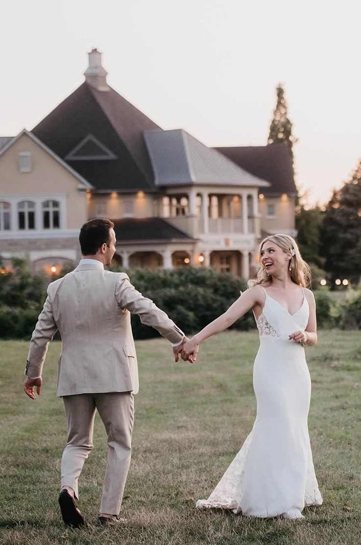 Bride in a V-neck satin gown with lace detail, is holding hands with groom in a tan suit outside a farmhouse as the sun sets. 