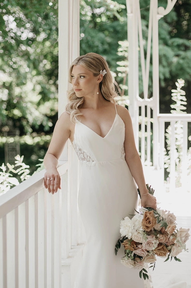 Bride in a satin bodice V-neck wedding dress with lace details and fit and flare skirt, is leaning on a white porch railing.