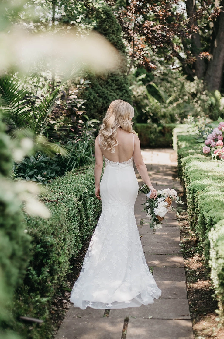 Back of bride in a lace and satin gown with a low back and thin straps, is walking through a garden path lined with hedges.