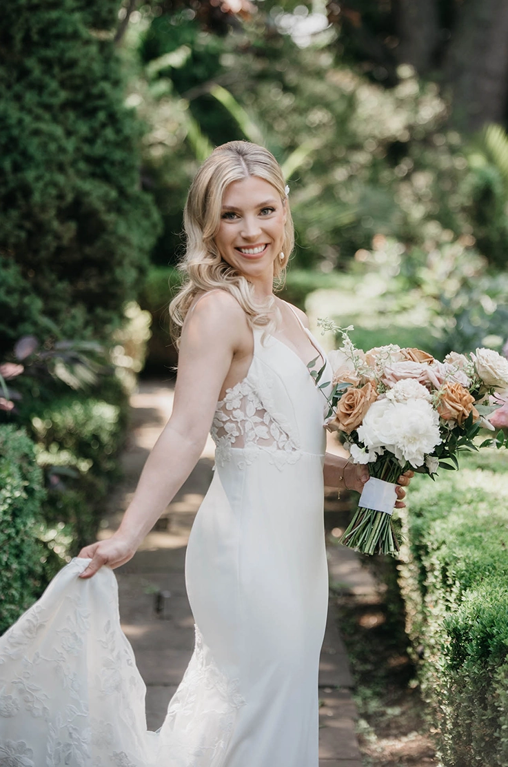 Close up of bride smiling in a satin fit and flare gown with lace side inserts, and is holding a bouquet on a sunlit path. 
