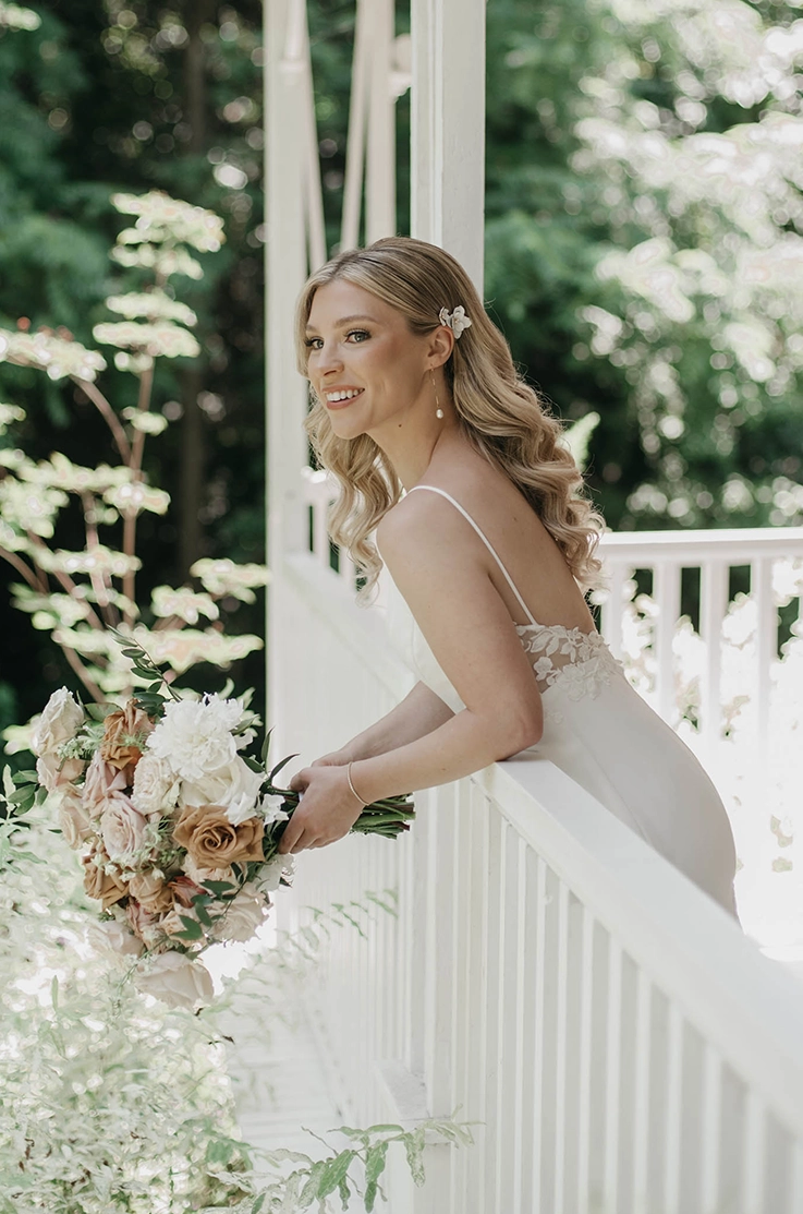 Bride leans on a white railing wearing a satin sleeveless gown with lace on the sides and is holding a bouquet outdoors. 
