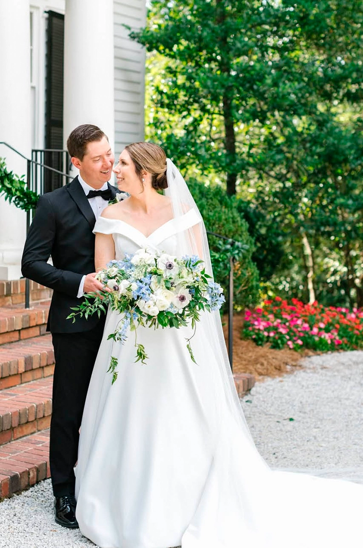 Bride in a portrait off-shoulder gown with an A-line satin faille skirt, poses with groom at the bottom of brick steps.