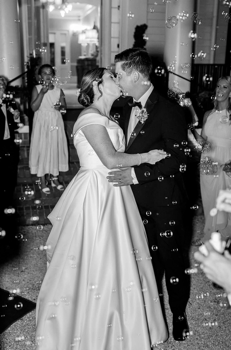 Bride in a portrait neck satin faille gown, kisses her groom near reception exit with bubbles floating in black and white.