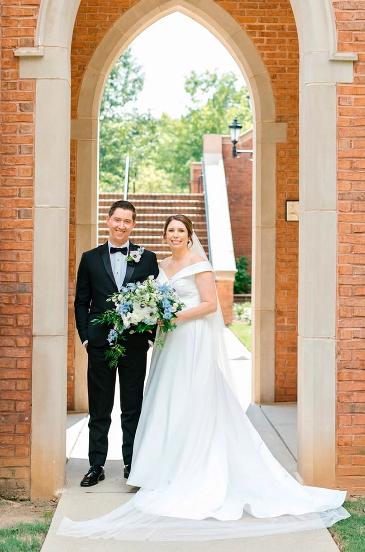Bride in a satin faille A-line gown with a portrait neckline and a veil, poses next to groom under a brick archway outdoors.
