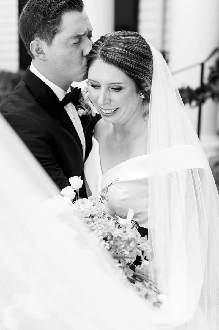 Close-up of groom embracing bride in a potrait off-shoulder gown with a long veil, and holds a bouquet outside the venue.