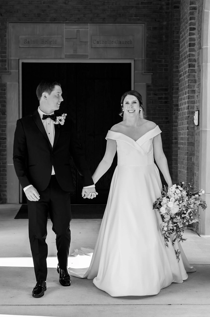 Bride in an off-shoulder A-line wedding dress, walks hand in hand with groom, outside a church entrance in black and white.