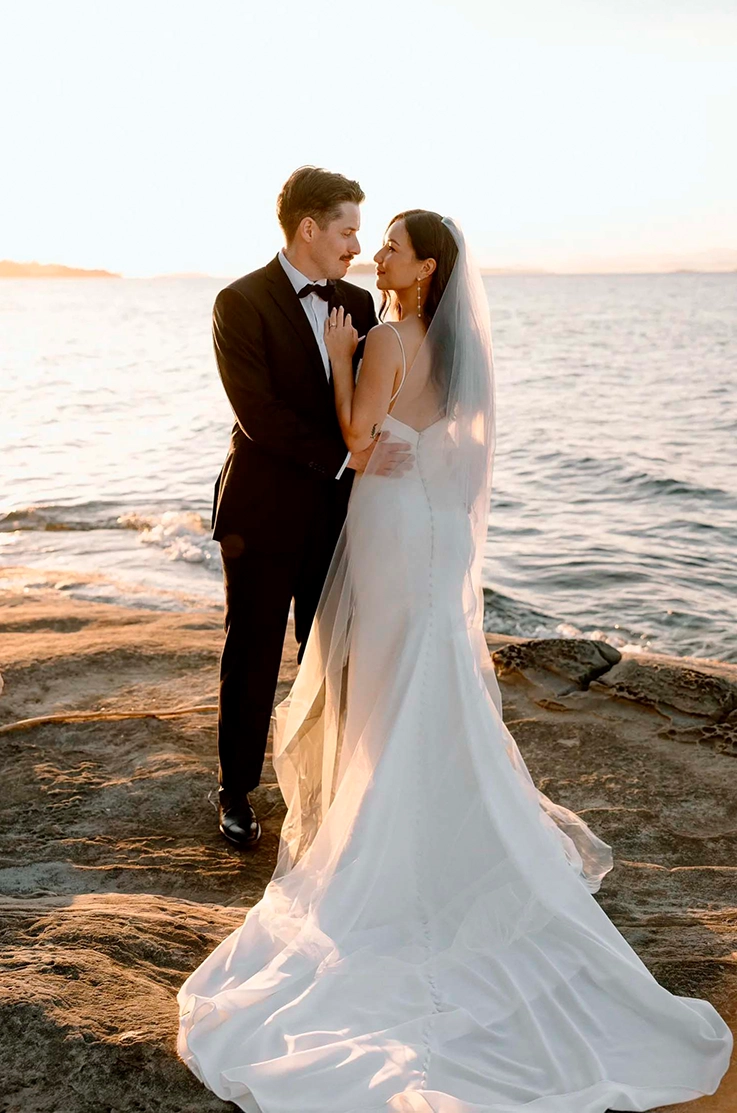 Groom embraces his bride in a crepe fit and flare gown with buttons and a veil, on a rocky shore at sunset.