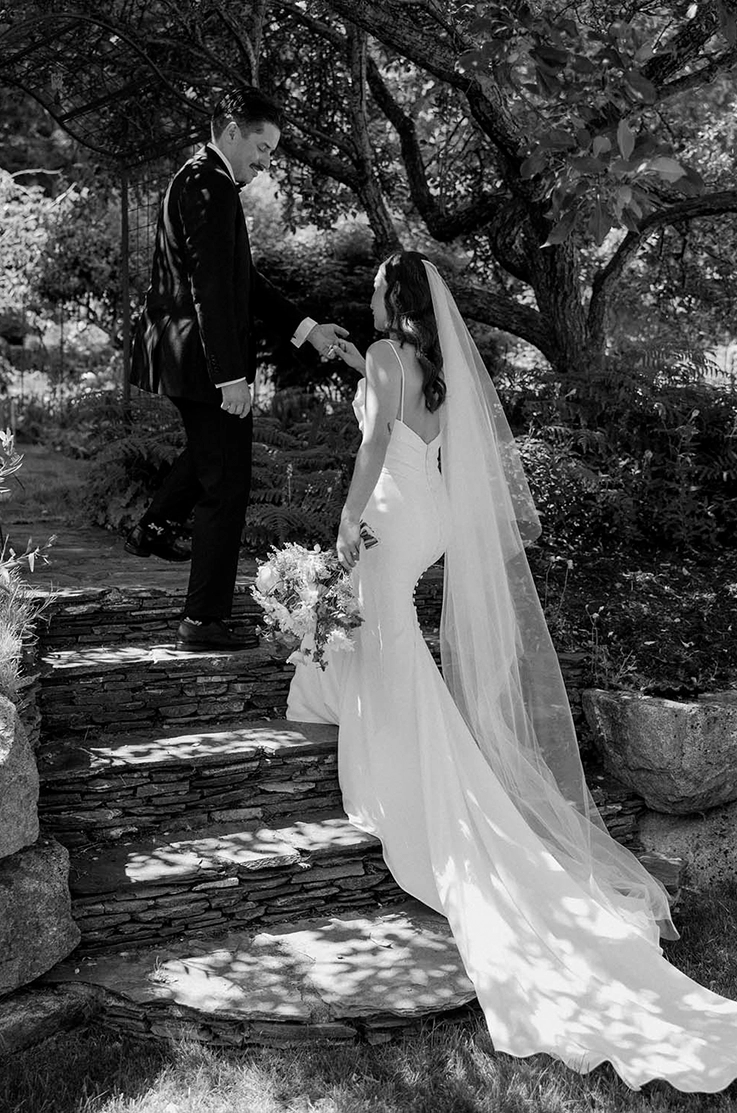 Bride in a low back crepe wedding dress with a long veil, walks up stone steps with her groom in black and white.
