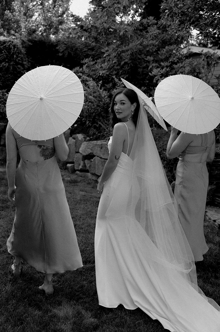 Bride looks over her shoulder in a crepe fitted gown with a low back and a veil, next to bridesmaids holding umbrellas.