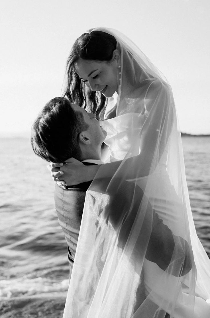 Groom lifts up his bride in a scoop neck crepe gown with pleating and a veil, in front of an ocean in black and white. 