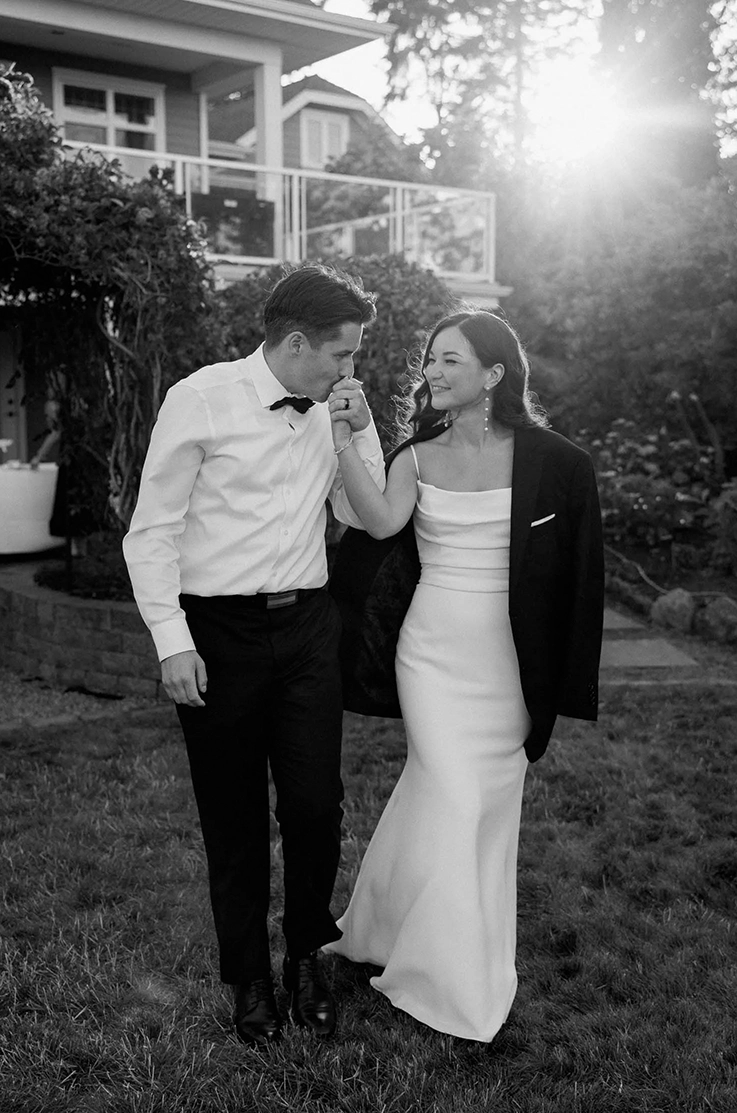 Bride in a crepe fit and flare gown with a pleated bodice walks alongside her groom, near the venue in black and white.