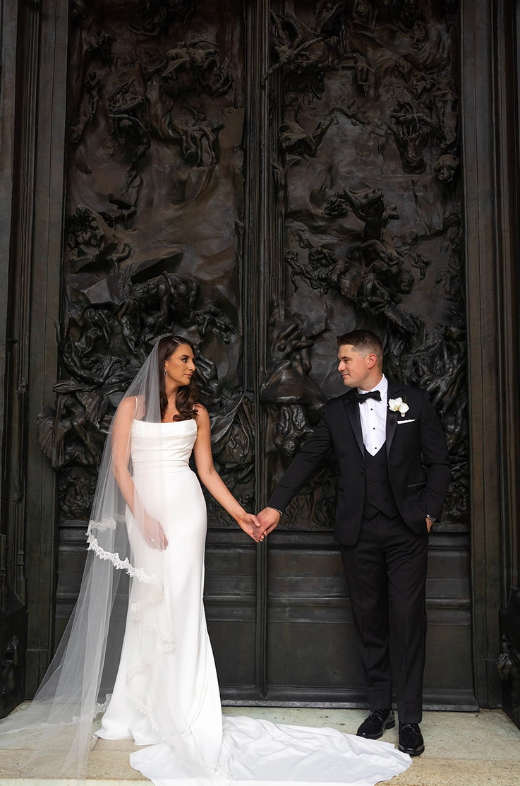 Bride in a crepe pleated bodice gown with fit and flare skirt and a lace-trim veil, poses with groom in front of large doors.
