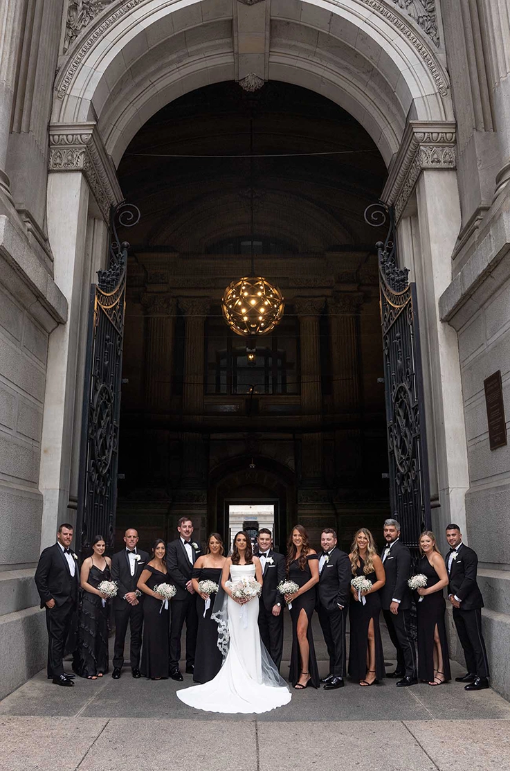 Bride in a crepe fit and flare gown with a lace-edge veil poses next to groom and the bridal party, at a grand building entrance