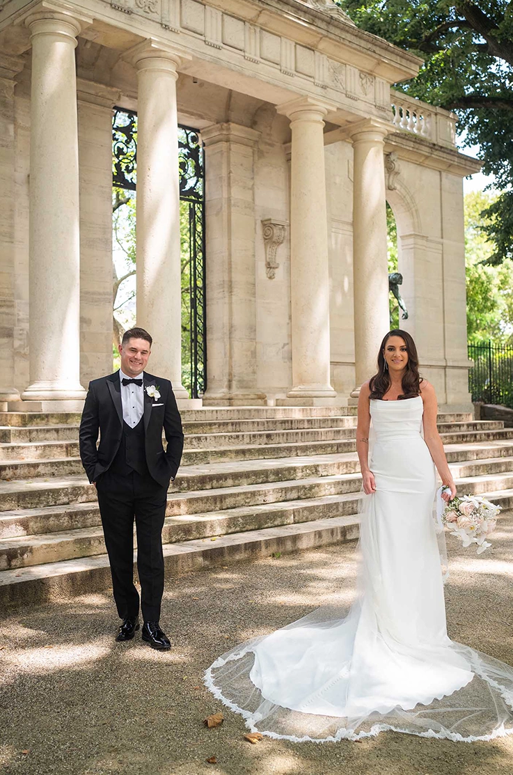 Bride in a crepe fit and flare gown with a pleated bodice and a veil, poses near groom in front of stone steps outdoors. 