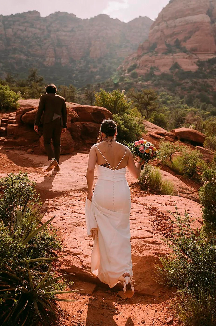 Bride in a low back fitted wedding dress with thin straps, holds her train and walks behind her groom in a red rock canyon.