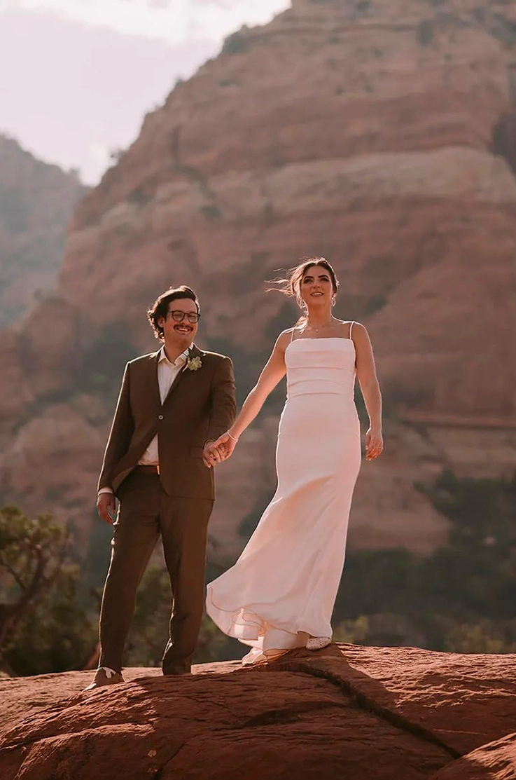 Bride in a crepe fit and flare gown with a pleated bodice and spaghetti straps, stands in a canyon, and poses with groom. 