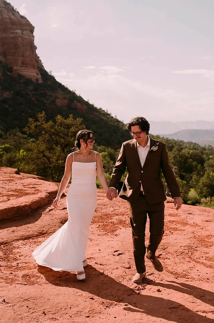 Bride in a fit and flare crepe gown with pleating and thin straps, walks hand in hand with her groom in a vast desert.
