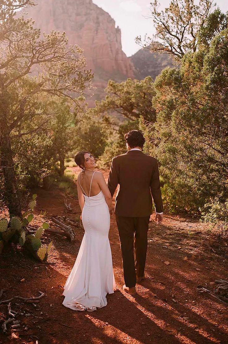 Bride looks over her shoulder in a crepe fit and flare gown with a low buttoned back, walking beside groom on a desert trail.
