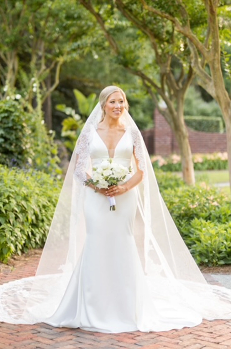 Bride in a plunging neck satin gown with fit and flare skirt and a long veil, holds a bouquet on a path lined with trees.